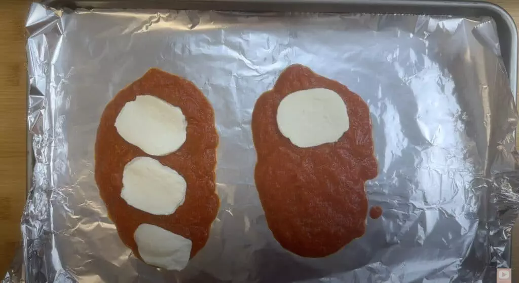 Two pieces of bread topped with tomato sauce and mozzarella on a foil-lined baking sheet.